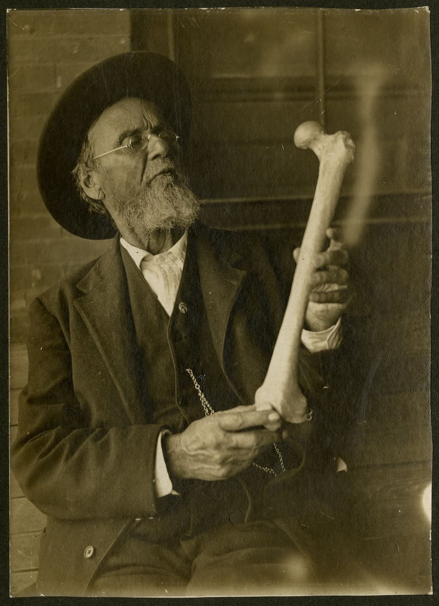 Andrew Taylor Still, an elderly bearded man in a hat and wool suit, closely studying a femur held in both hands