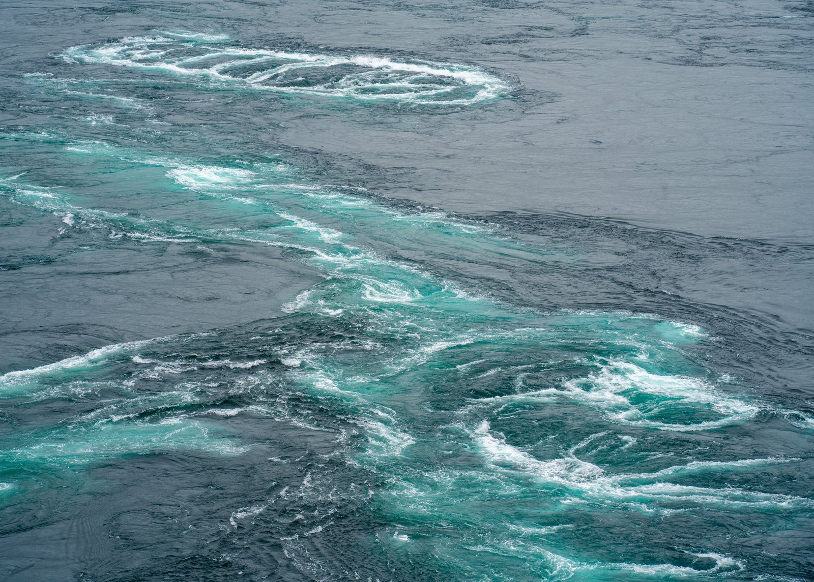 Tidal currents turning in spiral eddies of turquoise and white across slate-blue water
